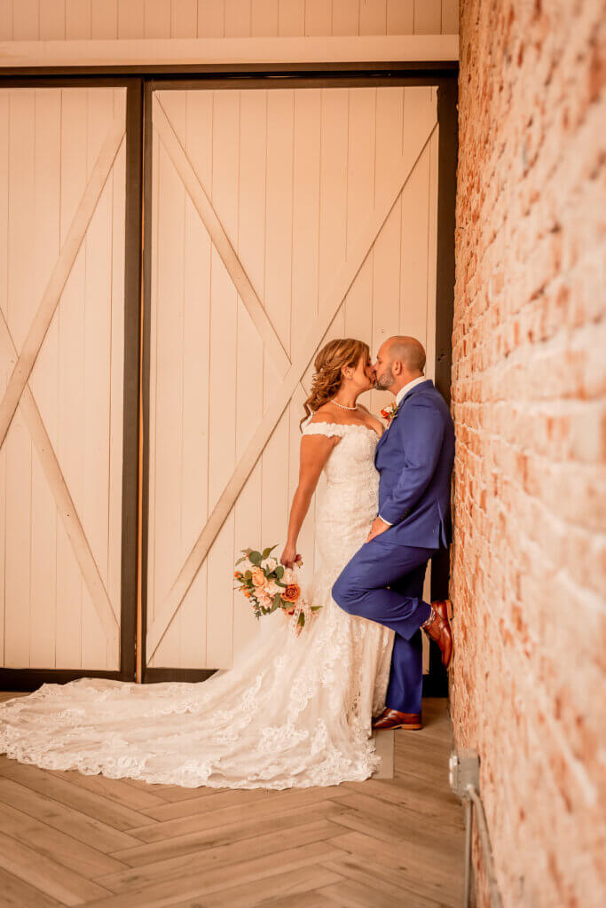 Photo of groom leaning up on red brick wall with bride kissing him and her bouquet of flowers hanging down
