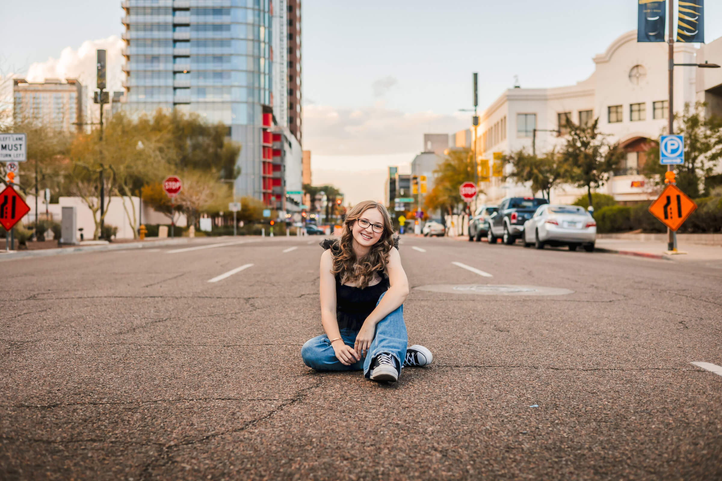photo of senior grad in downtown Phoenix sitting in the middle of the street with buildings and cars behind her in the background
