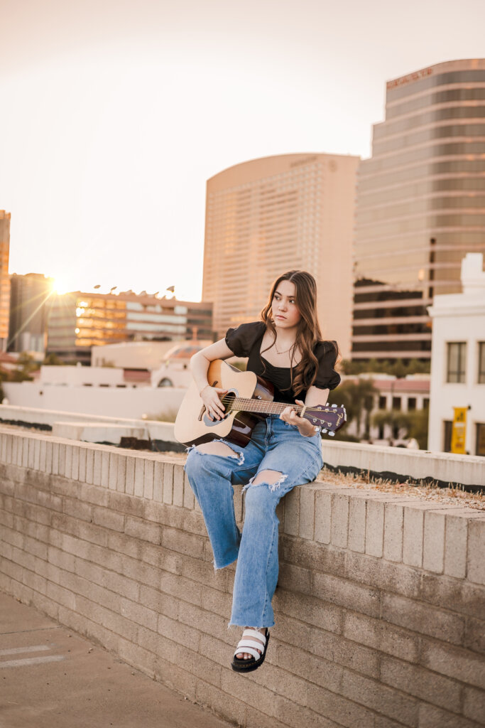 Senior girl sitting on the edge of a rooftop, strumming her guitar with Phoenix cityscape behind her.