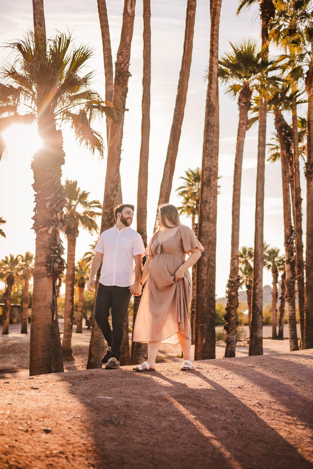 Pregnant woman holding hands with husband in nude dress by Palm Trees at Papago Park during golden hour in Phoenix, AZ.