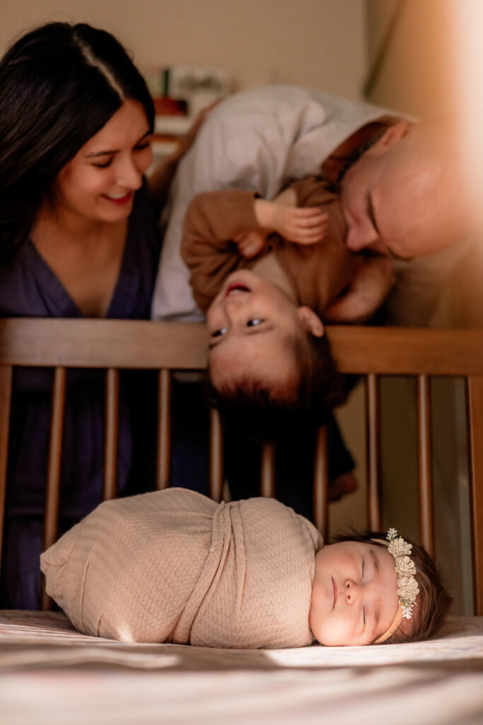 Parents and brother being tipped upside down in the background while focus is on new baby arrival during Phoenix In-Home Newborn Photography Session.