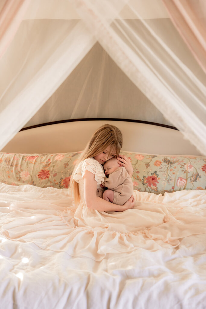 Older sister with flowy dress spread out on bed, holding new baby sister during newborn photoshoot.