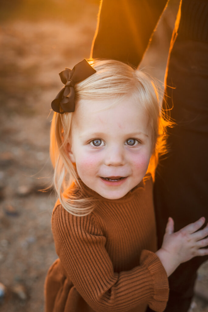 Close up photo of little girl holding on to dad's leg with golden sun hitting the back of her blonde hair.