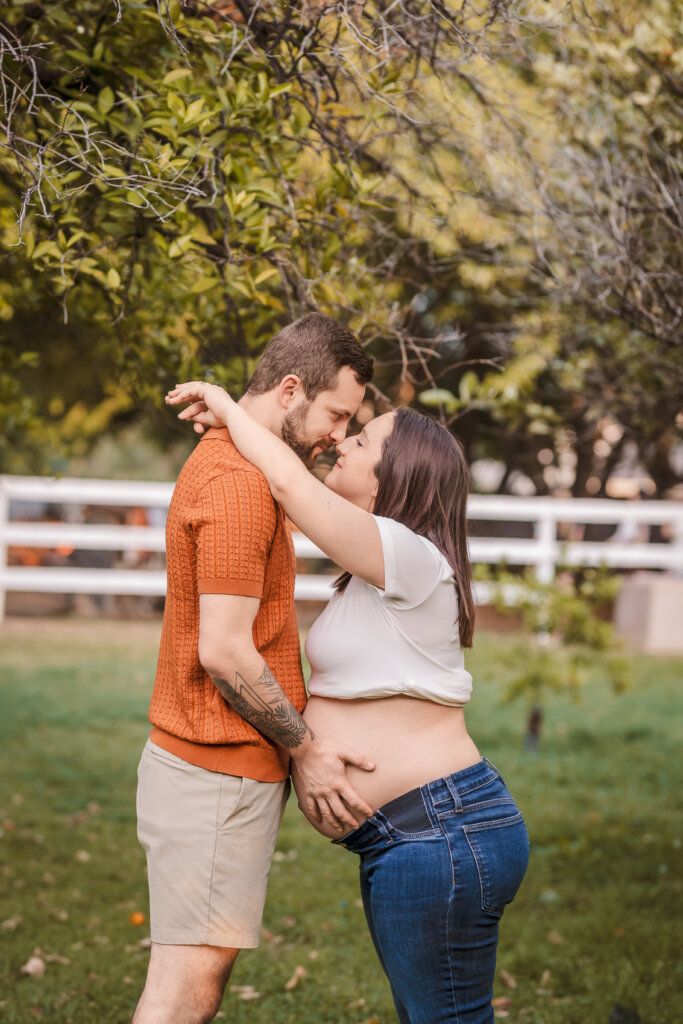 Phoenix maternity photographer captures cute moment of expecting couple leaned in together with hands on belly with white picket fence in the background at Manistee Ranch Park in Glendale, Arizona.