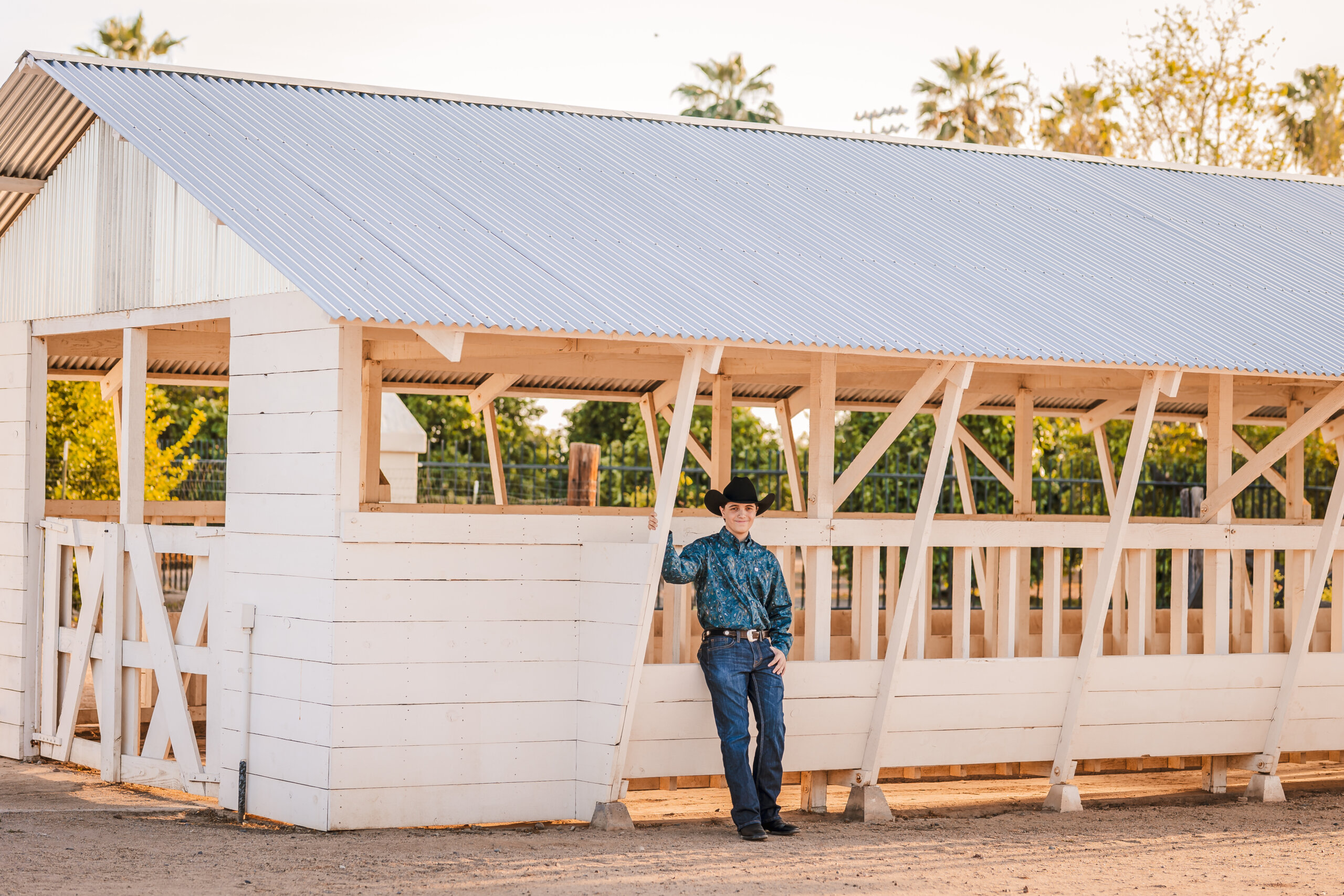Senior portrait with high school student dressed in western attire leaning up against white barn at Sahuaro Ranch Park in Glendale, Arizona.