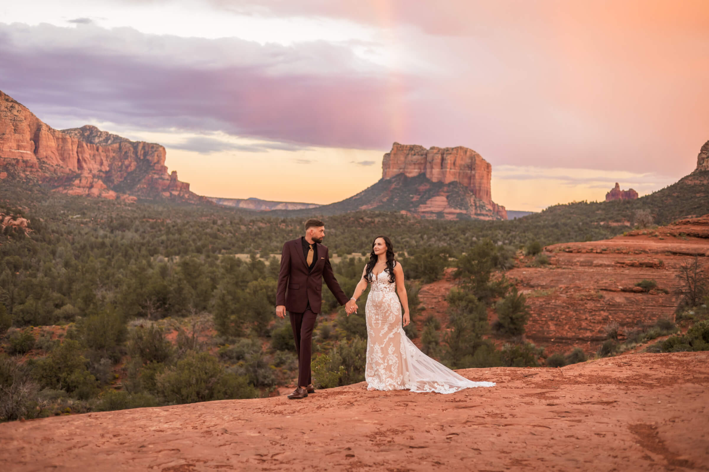 Bride and groom holding hands beneath colorful evening clouds at a Sedona overlook